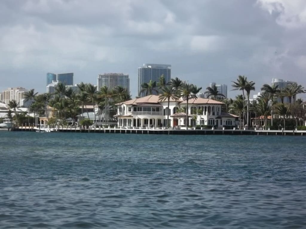 Waterfront mansion with palm trees and city skyline.